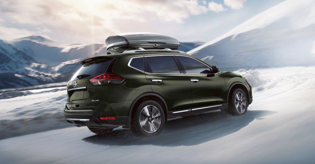 A metallic dark green 2017 Nissan Rogue driving down a snowy road with mountains in the background. There is cargo on the vehicle's roof rails.