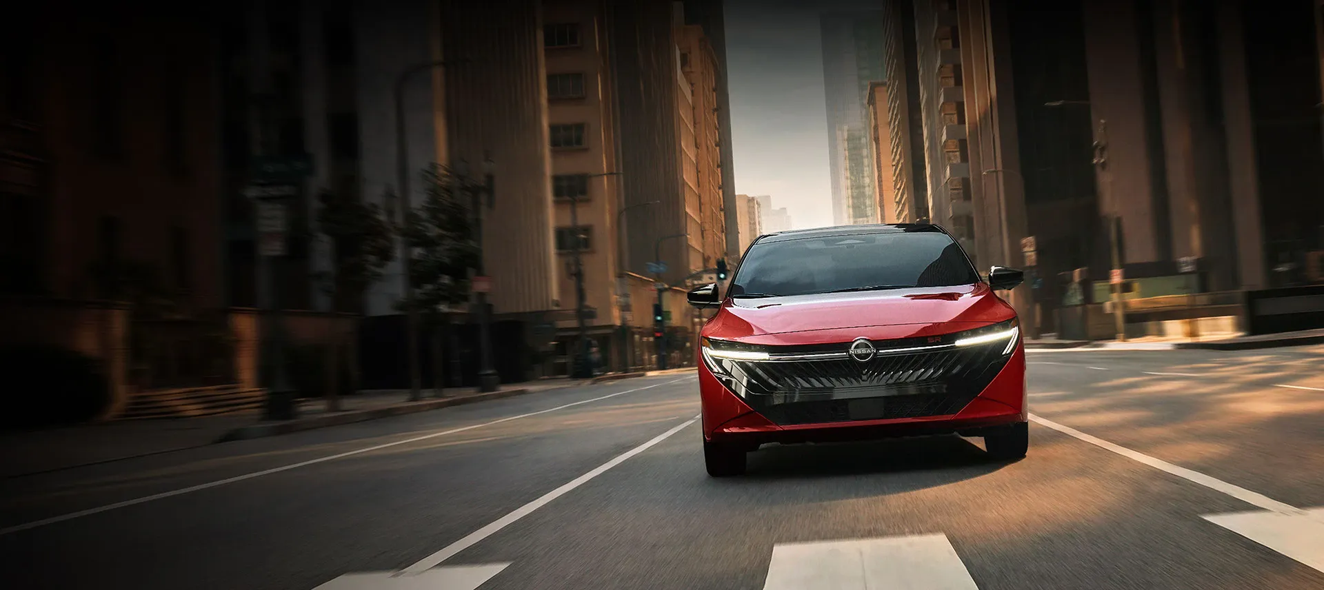 Front view of a red 2026 Nissan Sentra SR driving through downtown streets with LED headlights illuminated and a sporty black grille
