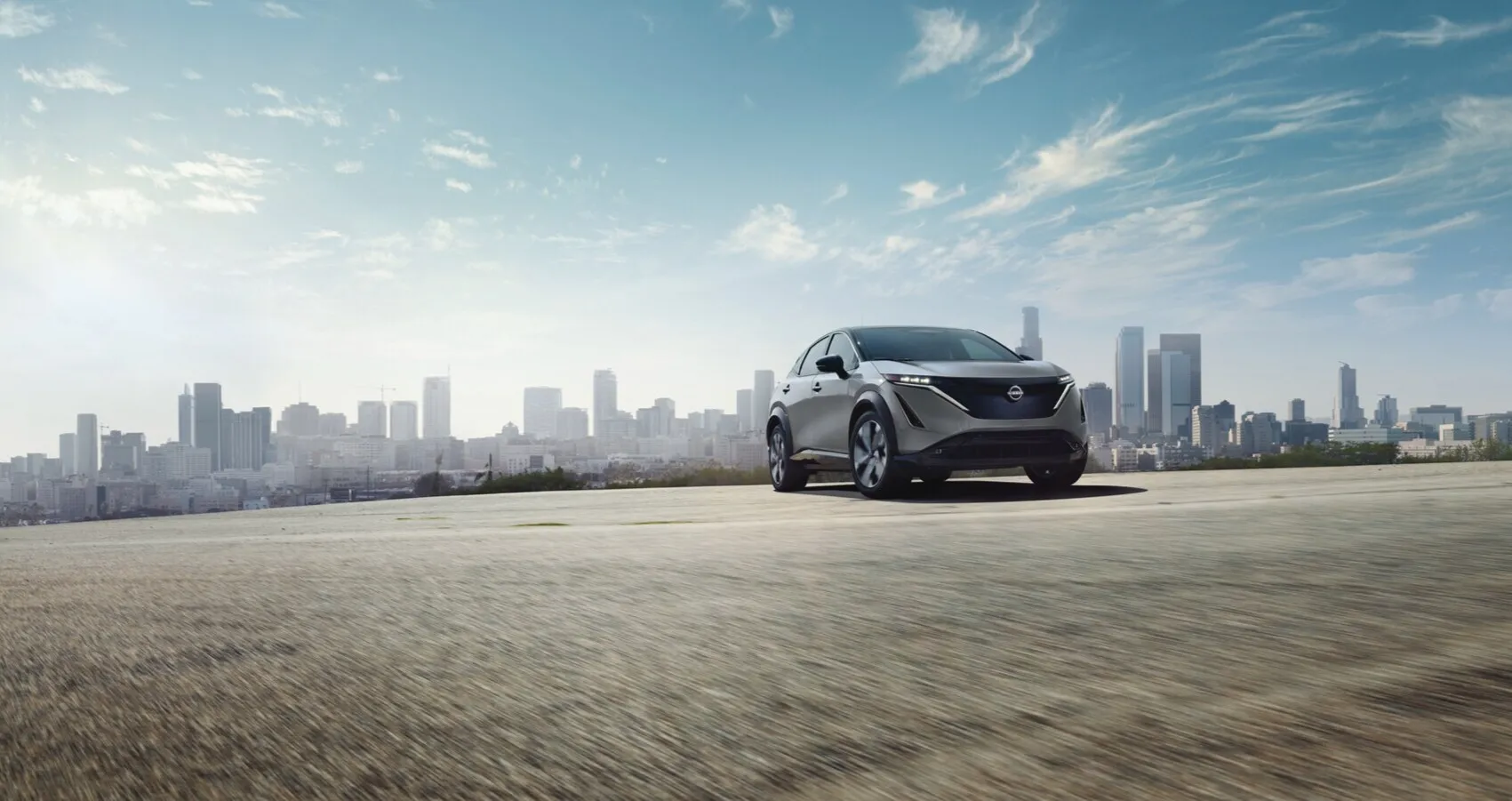 Silver Nissan Ariya parked on open concrete surface with city skyline in background